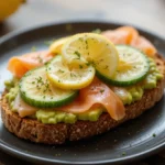 A slice of avocado toast topped with smoked salmon, cucumber, and a squeeze of lemon