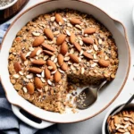 Baked oatmeal in a dish topped with sliced almonds, chia seeds, and a drizzle of almond butter