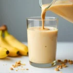 A peanut butter banana smoothie being poured from a blender into a glass, with a swirl of peanut butter on top